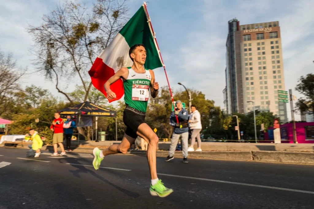 Un corredor del Maratón de la Ciudad de México avanza con determinación mientras sostiene una gran bandera de México. Viste un uniforme verde con la palabra “Ejército” y corre en una calle bordeada de árboles, con un edificio alto visible en el fondo. La escena captura la energía y el orgullo patriótico del corredor en una mañana soleada, mientras espectadores lo animan desde el costado.