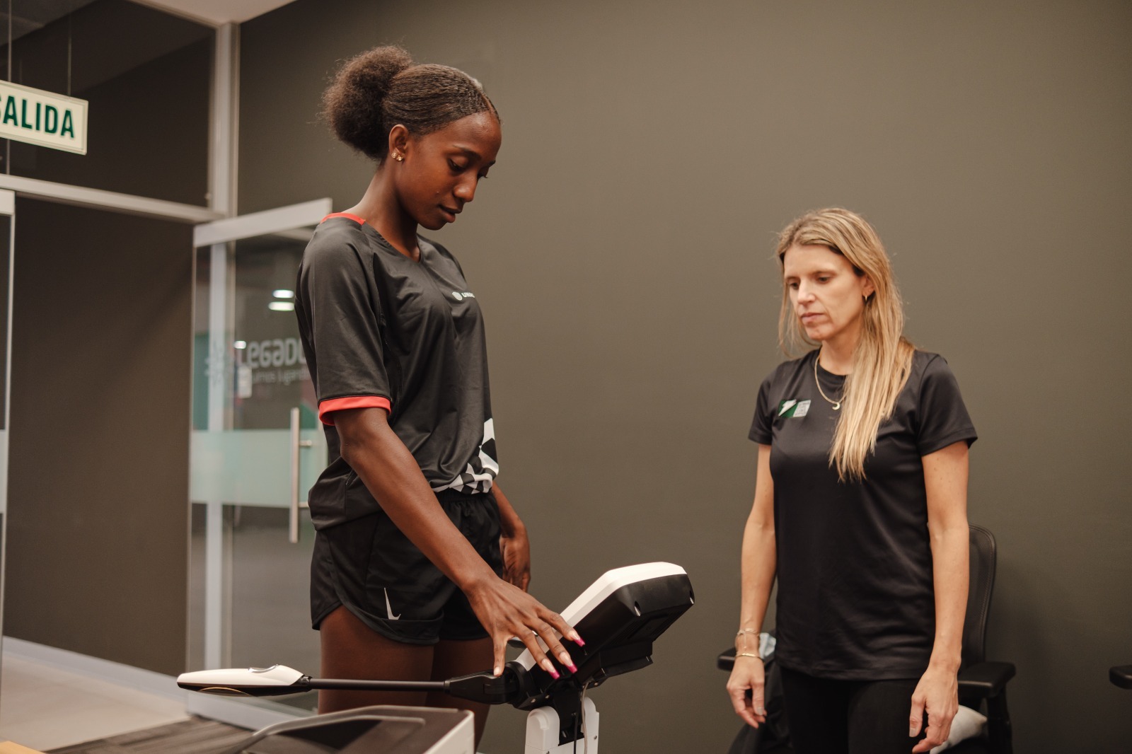 Una atleta se encuentra realizando una prueba de composición corporal bajo la supervisión de su nutricionista. La atleta, vestida con ropa deportiva negra, coloca su mano sobre un dispositivo de análisis mientras la entrenadora observa atentamente. Están en una sala moderna con paredes de color oscuro.