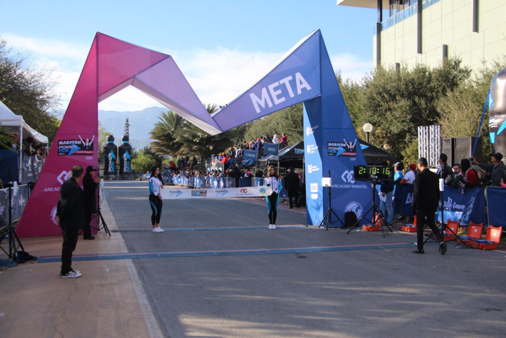 Un corredor de la Maratón de la Ciudad de México avanza con determinación mientras sostiene una gran bandera de México. Viste un uniforme verde con la palabra “Ejército” y corre en una calle bordeada de árboles, con un edificio alto visible en el fondo. La escena captura la energía y el orgullo patriótico del corredor en una mañana soleada, mientras espectadores lo animan desde el costado.