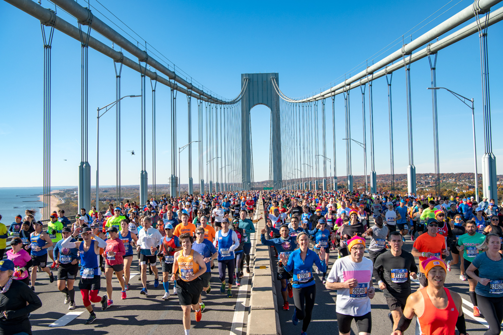 Una multitud de corredores participa en el Maratón de Nueva York, cruzando el imponente Puente Verrazzano-Narrows bajo un cielo azul claro. El paisaje urbano y costero se extiende a ambos lados, mientras los corredores avanzan con determinación, llenando toda la calzada del puente. El ambiente es enérgico y vibrante, reflejando la magnitud de este evento deportivo en una de las ciudades más icónicas del mundo.