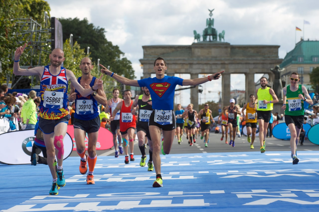 Corredores celebran al cruzar la línea de meta en el Maratón de Berlín, con la icónica Puerta de Brandeburgo al fondo. Uno de los participantes lleva una camiseta con el logo de Superman, levantando los brazos en señal de triunfo. El ambiente es de alegría y satisfacción mientras los competidores finalizan la carrera en una jornada soleada.